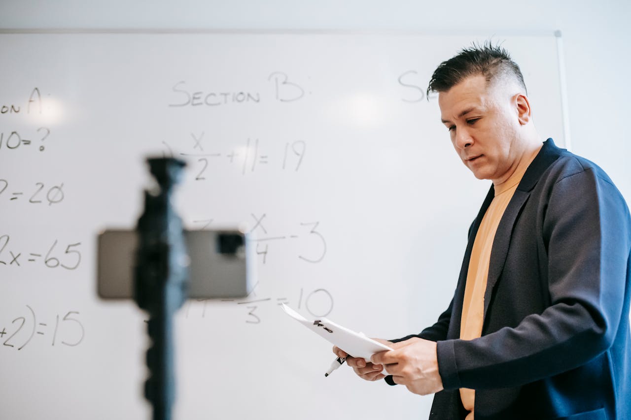 Educator teaching online math class, using whiteboard and digital device.