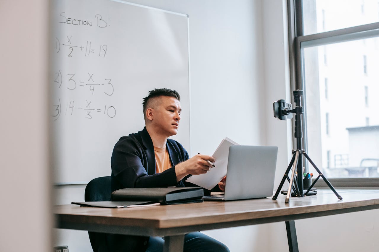 A male teacher conducting an online class with a laptop and camera setup in a classroom.